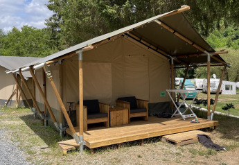 Tienda safari con terraza de madera y sillones exteriores, ubicada en un camping verde junto a árboles.