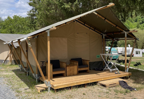 Tienda safari con terraza de madera y sillones exteriores, ubicada en un camping verde junto a árboles.