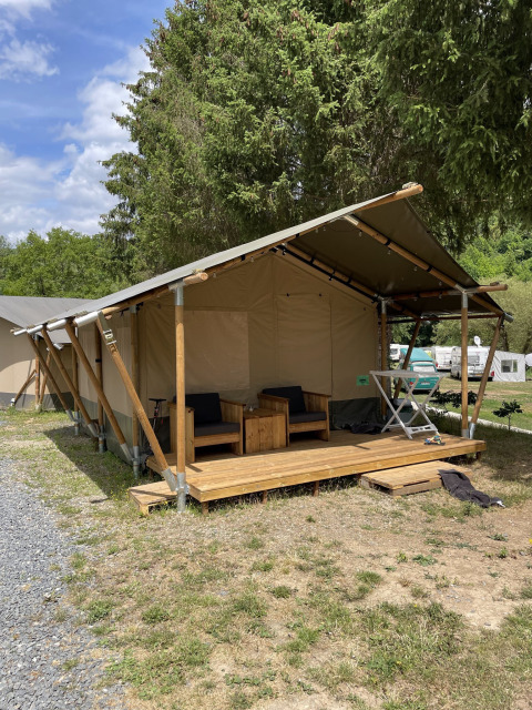 Tente safari avec véranda en bois et fauteuils extérieurs, installée sur un camping herbeux près des arbres.