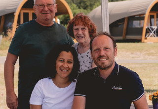 Four people smiling together at a picnic bench in front of glamping tents at Camping de Bovenberg, Overijssel.