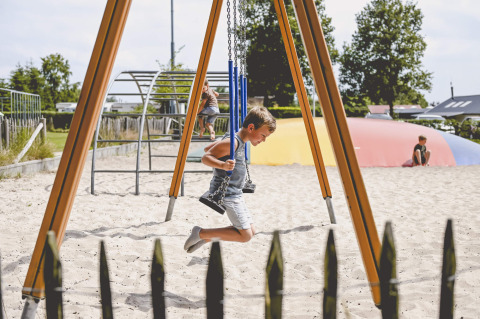 Ragazzo sull'altalena in un parco giochi sabbioso al Camping de Bovenberg, Overijssel, Olanda, giornata soleggiata.