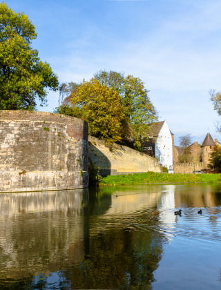 Blick auf eine steinerne Festungsmauer mit Teich, Enten und viel Grün in einer Ferienanlage.
