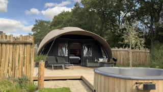 Modern tent-style lodge with deck, sun loungers, wooden hot tub, and wooden fence, surrounded by greenery.