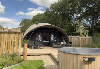En moderne teltlignende lodge med terrasse og boblebad i træ, omgivet af natur og træhegn under blå himmel.