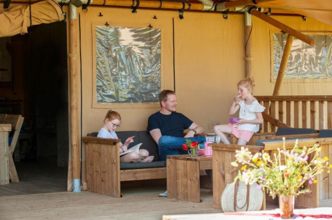 Gezin geniet van de veranda bij safaritent Meddo met houten tuinmeubelen en bloemen.
