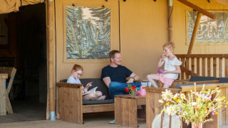 Gezin geniet van de veranda bij safaritent Meddo met houten tuinmeubelen en bloemen.