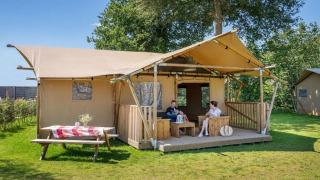 Twee personen zitten op de veranda van safaritent Hilgelo, omringd door natuur en zonlicht.