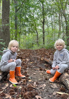 Twee kinderen in grijze kleding en oranje laarzen zitten op boomstammen in het bos bij Camping de Boskant.