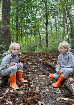 Zwei Kinder in grauer Kleidung und orangefarbenen Stiefeln sitzen im Wald im Ferienpark Camping de Boskant.