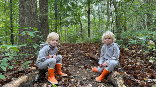 Zwei Kinder in grauer Kleidung und orangefarbenen Stiefeln sitzen im Wald im Ferienpark Camping de Boskant.