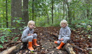 Dos niños con ropa gris y botas naranjas sentados en troncos en el bosque de Camping de Boskant en Limburgo.