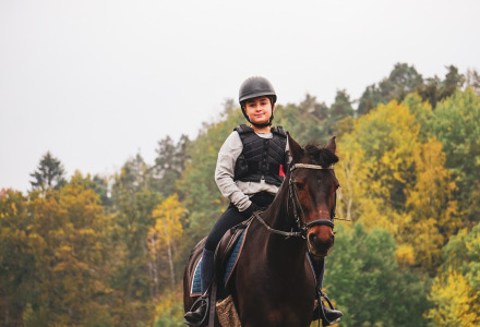 Kind met helm en vest rijdt op een paard voor herfstbomen in een vakantiepark met glamping.