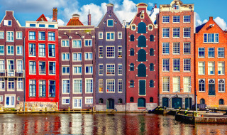 Fachadas coloridas de casas estrechas junto al canal en Ámsterdam, Países Bajos, bajo un cielo azul y nubes.