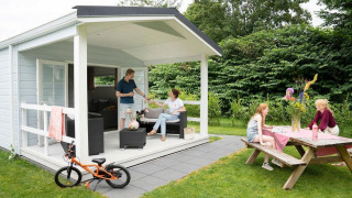 Gezin geniet van glamping aan een hutje met overdekt terras en picknickbank in een groene tuin.