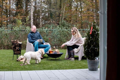 Couple sits by a fire pit with their dog and coffee outside a cabin at Camping Sevink Molen, Netherlands.