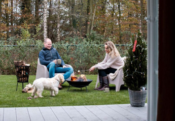 Coppia seduta vicino al fuoco con cane e caffè davanti a una baita al Camping Sevink Molen, Paesi Bassi.