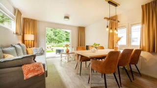 Lys og hyggelig stue med spisebord og sofa i et glampingophold, udsigt til have gennem glasdør.