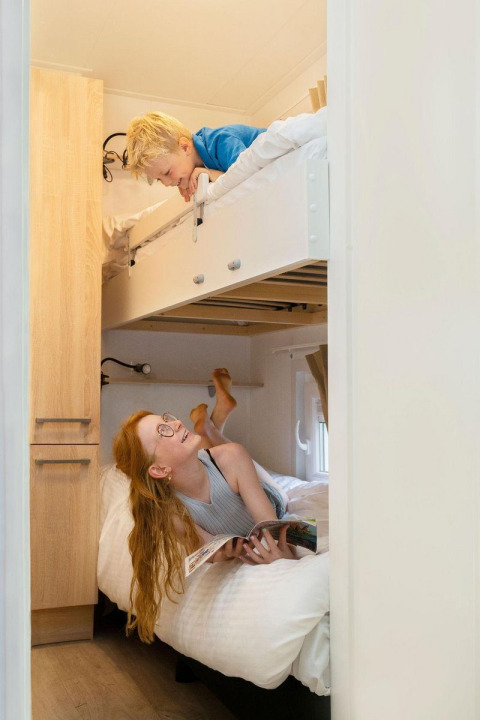 Dos niños disfrutan de una habitación glamping con literas; la niña lee abajo y el niño sonríe arriba.