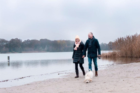 A couple walks with their dog on a lakeside beach by the Verandah Chalet at Camping Sevink Molen, Netherlands.