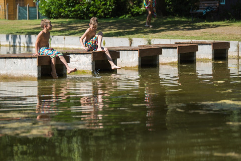 Zwei Kinder sitzen auf einem Holzsteg und planschen mit den Füßen im Wasser im Jocomo Parc, Belgien.
