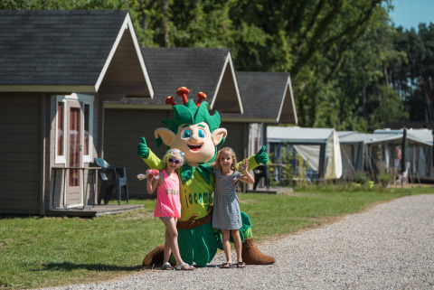 Zwei Kinder posieren mit einem Maskottchen vor Hütten im Jocomo Parc, einem Ferienpark in Limburg, Belgien.