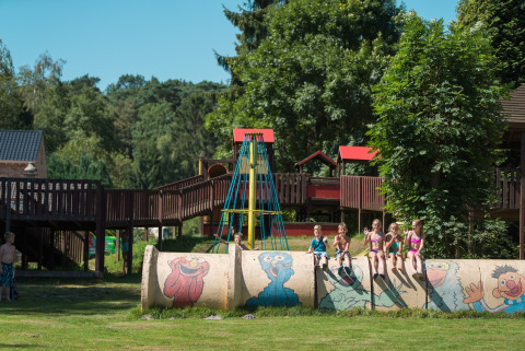 Des enfants jouent à Jocomo Parc, Belgique, assis sur un tuyau peint dans une aire de jeux verdoyante.