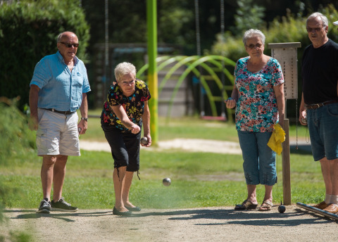 Cuatro adultos mayores juegan petanca al aire libre en un día soleado en Jocomo Parc, Limburg, Bélgica.