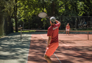 To personer spiller tennis på en solrig tennisbane omgivet af grønne træer i Jocomo Parc, Belgien.