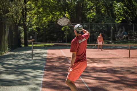 Twee mensen spelen tennis op een zonnige baan omgeven door bomen in Jocomo Parc, Belgisch Limburg.