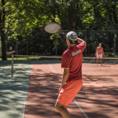 Dos personas juegan un partido de tenis en una pista soleada rodeada de árboles en Jocomo Parc, Bélgica.