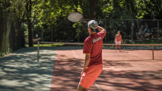 Dos personas juegan un partido de tenis en una pista soleada rodeada de árboles en Jocomo Parc, Bélgica.