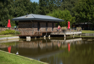 Een rustig gebouw aan het water in Jocomo Parc, Belgisch Limburg, met bomen en ontspannen bezoekers.