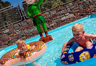 Zwei Kinder spielen in einem Pool mit Schwimmreifen, neben einer grünen Elfstatue im Jocomo Parc, Belgien.