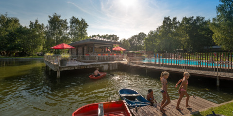 Niños juegan junto al lago y la piscina en el parque vacacional Jocomo Parc en Limburg, Bélgica, rodeados de naturaleza.