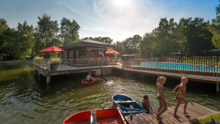 Niños juegan junto al lago y la piscina en el parque vacacional Jocomo Parc en Limburg, Bélgica, rodeados de naturaleza.