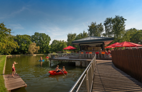 Bambini giocano sull’acqua e persone si rilassano al bar con ombrelloni rossi nel Jocomo Parc, Belgio.