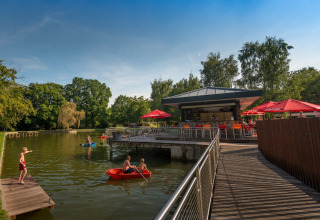 Bambini giocano sull’acqua e persone si rilassano al bar con ombrelloni rossi nel Jocomo Parc, Belgio.