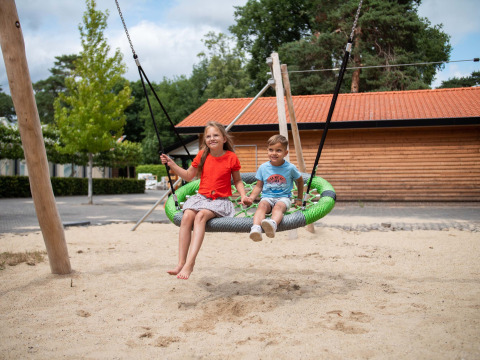 Two kids enjoying a large nest swing in the sandy playground at Resort Veluwe holiday park, Gelderland.
