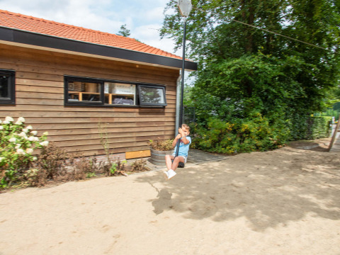 Niño juega en una tirolina en Resort Veluwe, parque vacacional en Gelderland, Países Bajos, junto a edificio y árboles.