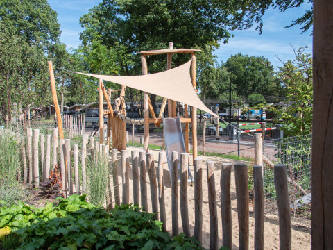 Playground with wooden fence, sand, and shade sail at Bospark Ede, a holiday park in Gelderland, Netherlands.