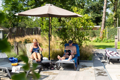 Twee mensen relaxen op ligstoelen onder een parasol in Bospark Ede, een vakantiepark in Gelderland, Nederland.