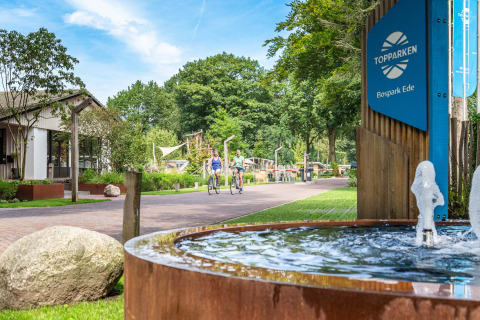 Parque de vacaciones Bospark Ede en Gelderland, Países Bajos, con ciclistas y fuente en la entrada soleada.