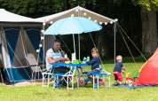 En familie camperer og spiser ved et bord under en parasol på Camping La Colline i Belgien Luxembourg.