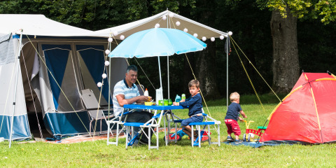 En familie camperer og spiser ved et bord under en parasol på Camping La Colline i Belgien Luxembourg.