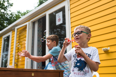 Deux garçons soufflent des bulles de savon devant un chalet jaune au Camping La Colline, Belgique Luxembourg.