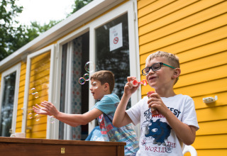 Due bambini soffiano bolle di sapone davanti a una casetta gialla a Camping La Colline, Belgio Lussemburgo.