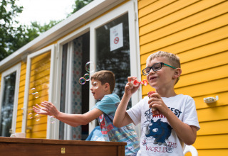 To drenge blæser sæbebobler foran et lyst gult sommerhus på Camping La Colline i Belgien Luxembourg.