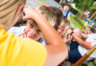 A child gets face painting at Camping La Colline, Belgium, watched by other children relaxing outdoors.