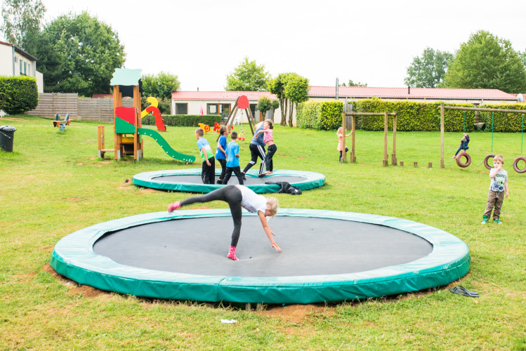 Kinder spielen auf Bodentrampolinen und einem Spielplatz im Camping La Colline, Belgien, mit viel Grünfläche.