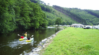 Camping la Roche en Bélgica mostrando personas en kayak en el río y caravanas al fondo bajo colinas verdes.